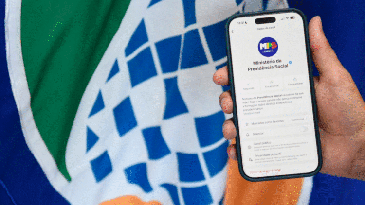 Mão segurando smartphone exibindo o canal oficial do Ministério da Previdência Social no WhatsApp, com logotipo ao fundo.