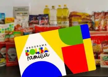 Cartão do Programa Bolsa Família em primeiro plano, com alimentos como leite, óleo e macarrão em segundo plano.