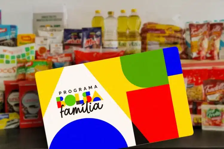 Cartão do Programa Bolsa Família em primeiro plano, com alimentos como leite, óleo e macarrão em segundo plano.