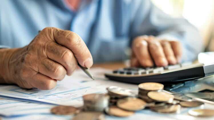 Idoso calcula aposentadoria com moedas, papéis e calculadora sobre a mesa.