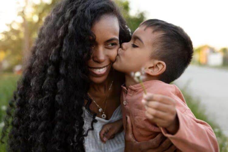 Criança dá beijo carinhoso na mãe sorridente em momento especial ao ar livre.