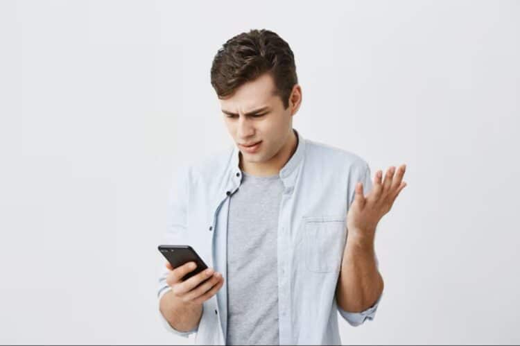 Homem jovem com expressão confusa e gesticulando com a mão, olhando para o celular, com fundo branco. Este texto é crucial para a acessibilidade, descrevendo a imagem para pessoas com deficiência visual e ajudando o Google a entender o conteúdo visual.