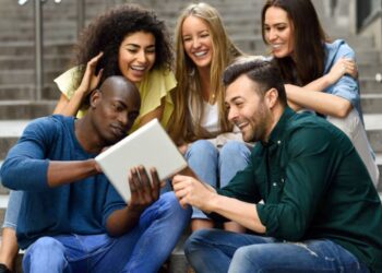 Grupo de jovens estudantes diversos e sorrindo, interagindo com um tablet em um ambiente urbano.