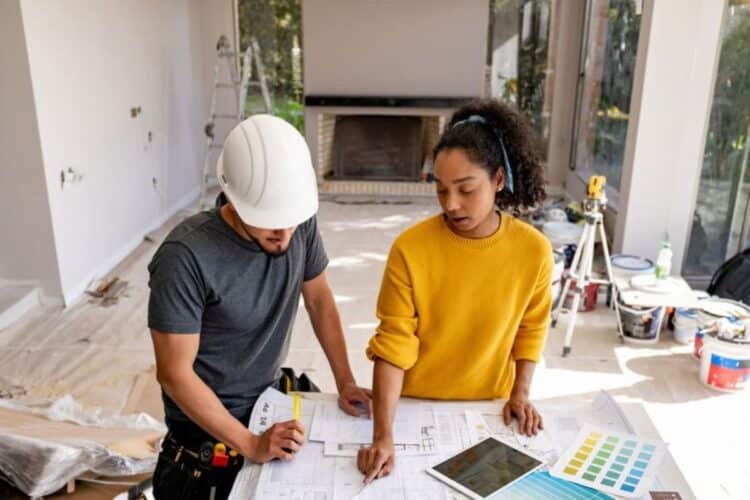 Um trabalhador da construção civil de capacete e uma proprietária analisando uma planta baixa de reforma. A imagem representa o planejamento para o programa de crédito "Reforma Casa Brasil".