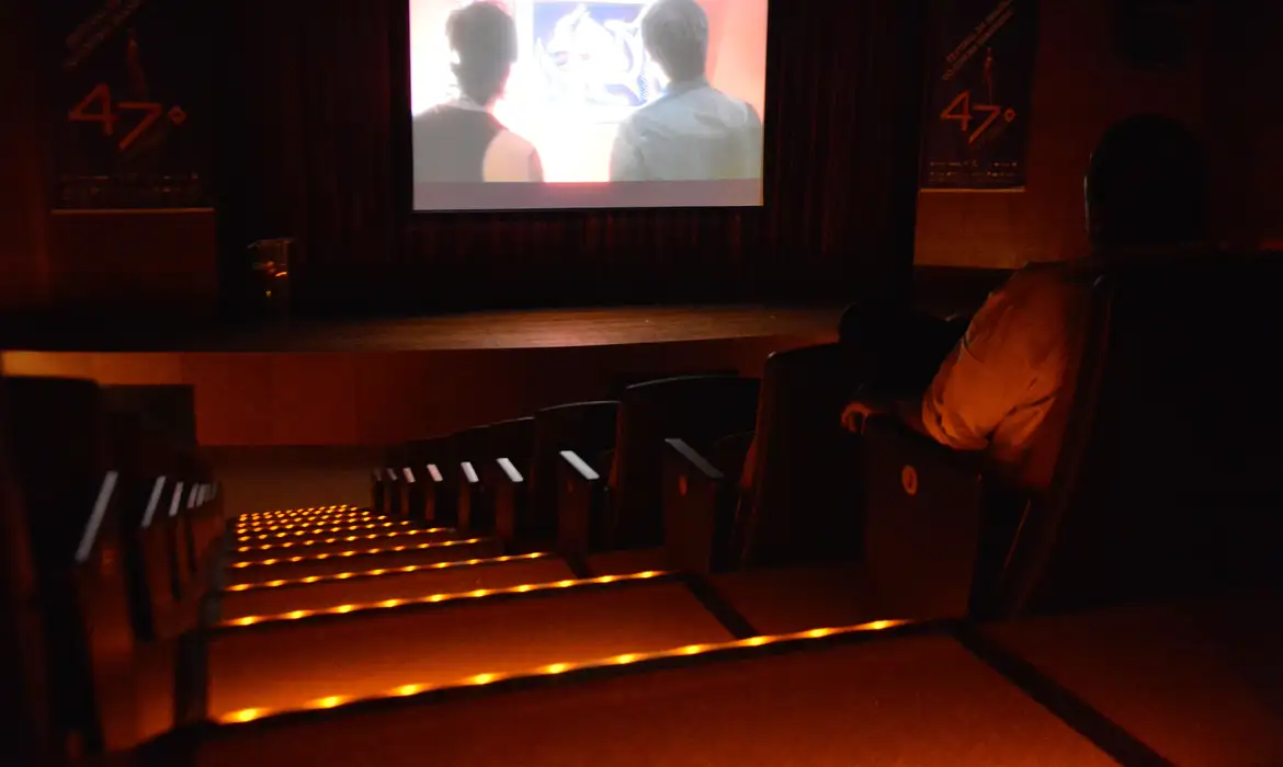 Documentacao-Meia-Entrada-Comprovacao-Beneficio-Cultural Interior de um auditório ou sala de cinema escurecida com poltronas vazias e iluminação de segurança nas escadas, representando o local onde o documento de meia-entrada (como o ID Jovem ou carteira de estudante) deve ser apresentado.