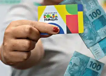 Mão segurando o cartão do Bolsa Família com notas de cem reais ao fundo, ilustrando a unificação dos pagamentos em estados específicos.