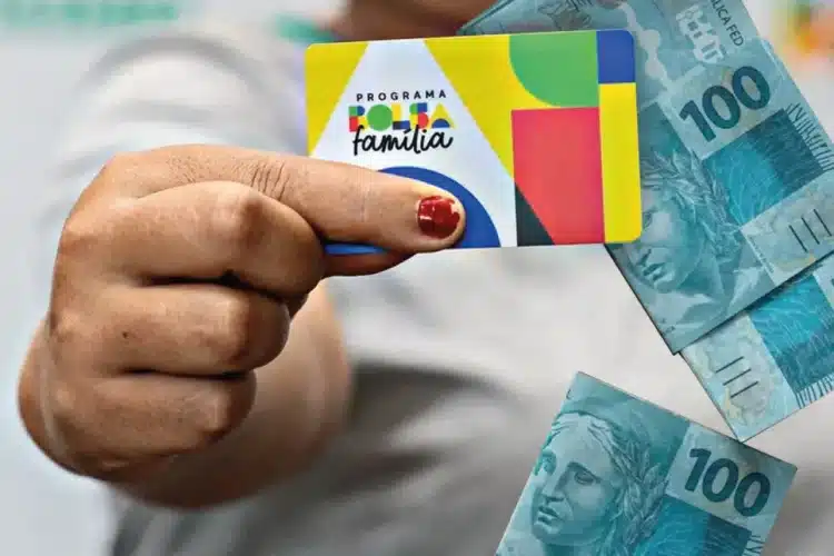 Mão segurando cartão do Programa Bolsa Família próximo a cédulas de dinheiro brasileiro
