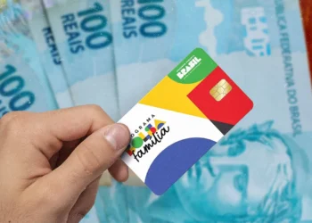 Mão segurando cartão do programa Bolsa Família com notas de 100 reais ao fundo