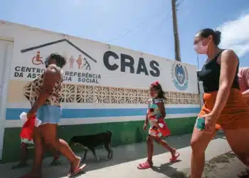 Urgente: 3 grupos podem ficar sem visita do CRAS — veja se você está na lista! 9 Pessoas, incluindo uma criança, caminhando em frente a um prédio com o letreiro 'CRAS - Centro de Referência de Assistência Social', indicando a busca por serviços sociais.