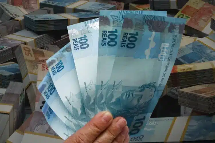 Pagamento extra de R$ 1.518,00 em outubro para CPFs com finais 3, 7, 0, 5, 2, 9, 1, 6, 8 e 4 1 Notas de 100 reais em mãos e empilhadas, simbolizando pagamento extra em outubro