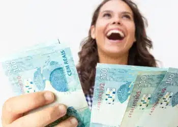 Mulher sorrindo e segurando várias notas de R$ 100, representando a alegria de receber o PIX ou pagamento na conta nesta quarta-feira (15).