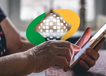 Mãos de idoso segurando e usando um smartphone em ambiente interno com logo da Previdência Social