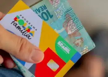 Cartão do Programa Bolsa Família com cédula de 100 reais sendo segurados por uma mão.