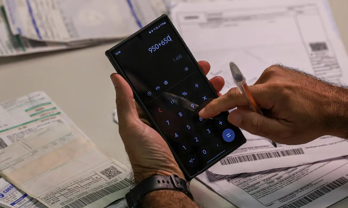 Cálculo de contas usando calculadora no celular Pessoa usando calculadora no celular para somar valores com várias contas sobre a mesa