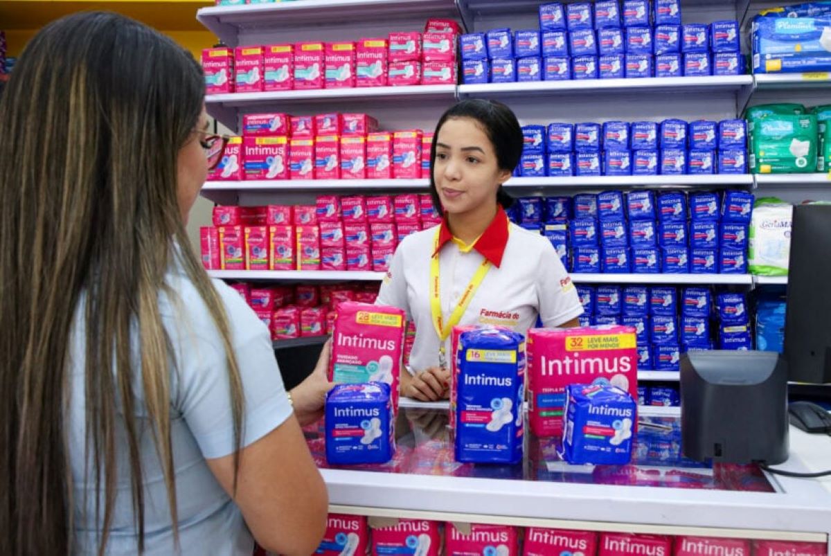 Atendente de farmácia auxiliando cliente em balcão com diversos pacotes de absorventes, indicando locais de distribuição gratuita.