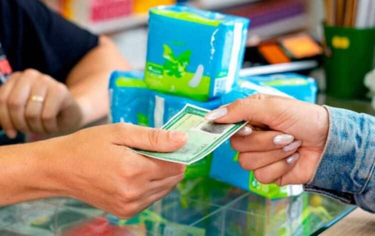 Pessoa entregando receita médica em uma farmácia para receber absorventes femininos gratuitos, com pacotes de absorventes ao fundo.