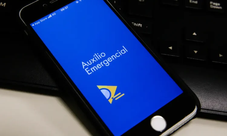 ATENÇÃO: Governo reabre prazo para devolver Auxílio Emergencial recebido indevidamente 1 Tela de smartphone mostrando o aplicativo Auxílio Emergencial aberto em fundo azul com símbolo amarelo