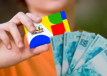 Pessoa segurando o cartão do Bolsa Família ao lado de notas de 100 reais, representando o recebimento do benefício.