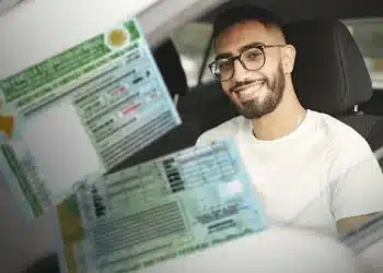 Homem sorridente vestindo camiseta branca mostrando carteira de motorista brasileira dentro do carro