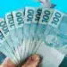 Bolsa Família, INSS, saque-aniversário e Gás do Povo: veja calendários de pagamentos para novembro e dezembro 11 Mão segurando várias notas de R$100 com outras notas caindo ao fundo, simbolizando os pagamentos do Bolsa Família, INSS, saque-aniversário e Gás do Povo em novembro e dezembro.