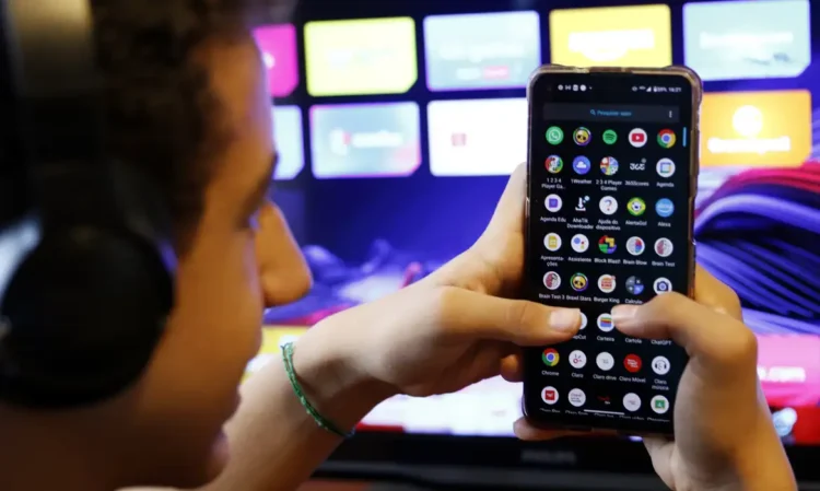 Pessoa usando smartphone com vários aplicativos visíveis na tela, ao fundo uma TV com interface colorida.