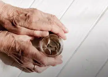 Mãos de idoso segurando um pote transparente com moedas de euro sobre mesa branca