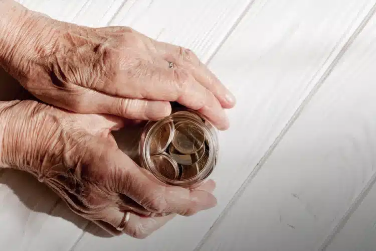 Três contas que idosos podem deixar de pagar no Brasil: confira seus direitos 1 Mãos de idoso segurando um pote transparente com moedas de euro sobre mesa branca