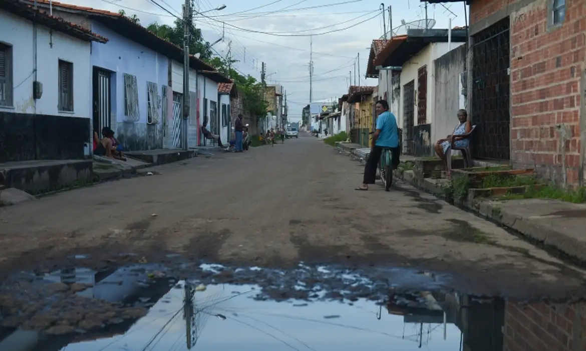 Rua residencial com casas simples e lamaçal Rua de terra com lama e poça d'água, casas modestas nas laterais, pessoas sentadas e homem com bicicleta
