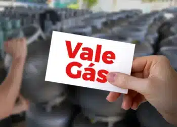 Mão segurando cartão "Vale Gás" com botijões de gás borrados ao fundo, simbolizando o benefício.
