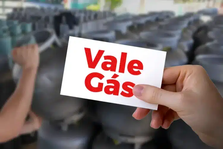 Mão segurando cartão "Vale Gás" com botijões de gás borrados ao fundo, simbolizando o benefício.