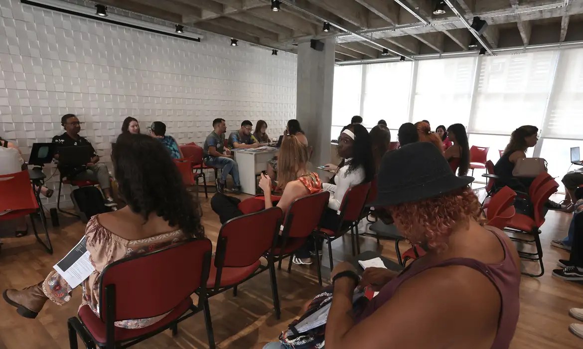 Evento com diversidade de participantes em ambiente moderno Grupo de pessoas sentadas em cadeiras vermelhas em sala iluminada, interagindo e lendo materiais