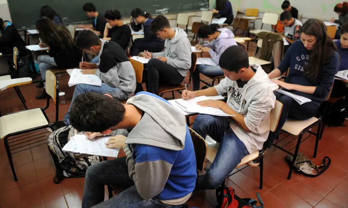 Alunos concentrados fazendo prova em sala de aula Estudantes sentados em carteiras escrevendo provas em uma sala de aula com chão de cerâmica
