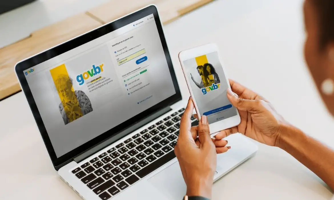 Login gov.br no computador e celular Pessoa usando celular e laptop com telas de login do gov.br em ambiente claro.