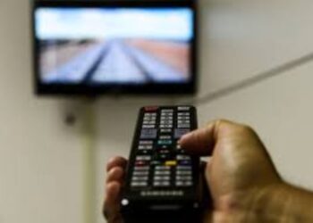 Mão segurando um controle remoto de TV, com a televisão desfocada ao fundo, simbolizando o uso de dispositivos como o 'gatonet' e o alerta urgente para seus usuários.