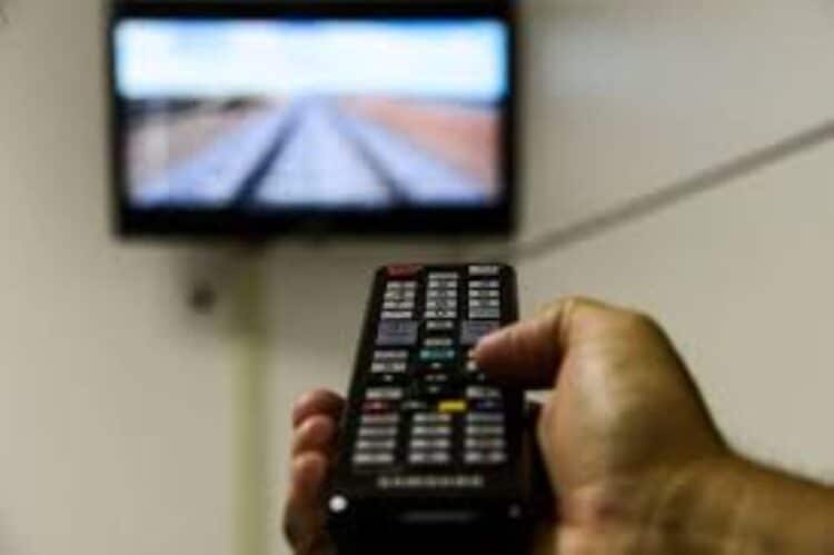 Mão segurando um controle remoto de TV, com a televisão desfocada ao fundo, simbolizando o uso de dispositivos como o 'gatonet' e o alerta urgente para seus usuários.