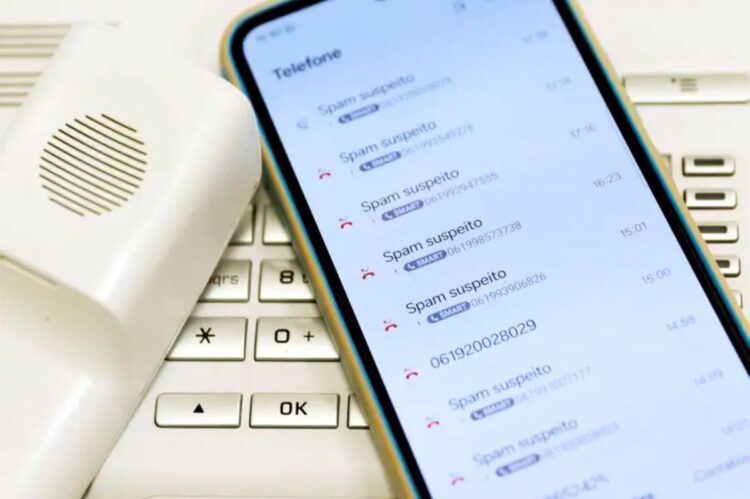 Tela de celular mostrando várias chamadas identificadas como spam suspeito ao lado de telefone fixo branco.