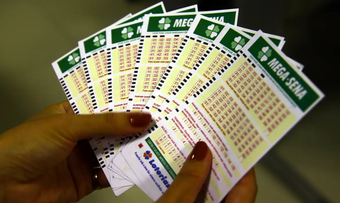 Bilhetes da Mega-Sena para aposta da loteria Mãos segurando vários bilhetes preenchidos da Mega-Sena para aposta na loteria federal