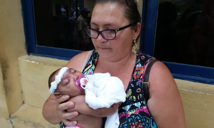 Mulher de óculos segurando bebê dormindo com faixa branca na cabeça e chupeta roxa