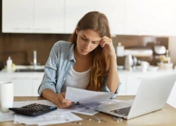 Mulher com expressão de preocupação em uma cozinha, revisando documentos e contas ao lado de uma calculadora e um notebook. A imagem ilustra a situação de quem busca o Feirão Limpa Nome para negociar dívidas e conseguir descontos nas finanças.