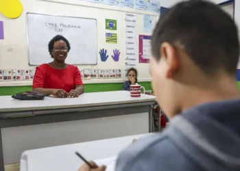 Professora sorridente sentada em sua mesa em sala de aula com alunos ao fundo escrevendo em cadernos
