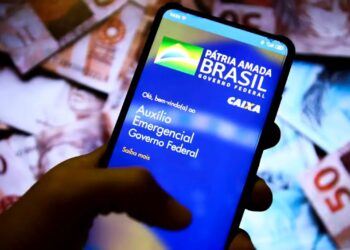 Mão segurando smartphone com o aplicativo "Auxílio Emergencial" na tela, notas de R$ 50 ao fundo, simbolizando a devolução de débitos.