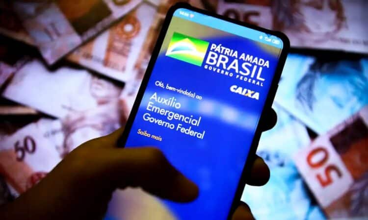 Mão segurando smartphone com o aplicativo "Auxílio Emergencial" na tela, notas de R$ 50 ao fundo, simbolizando a devolução de débitos.