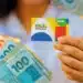 Confira as cidades com pagamento adiantado do Bolsa Família em dezembro 11 Pessoa segurando cartão do Programa Bolsa Família com cédulas de real em destaque