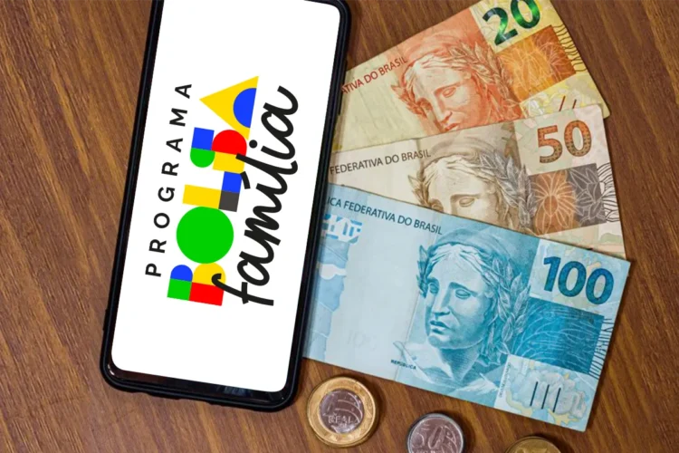 Celular mostrando logo do Programa Bolsa Família ao lado de cédulas e moedas de reais sobre mesa de madeira