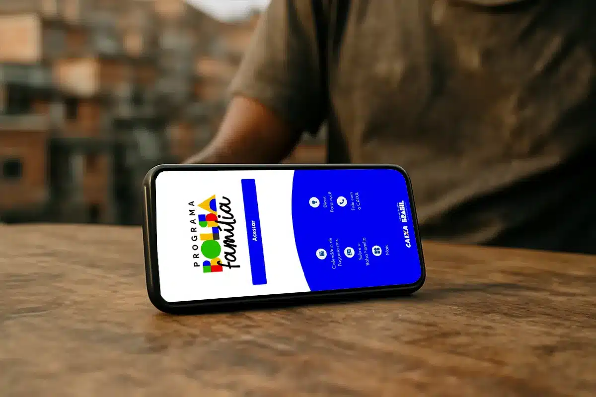 consulta digital calendario bolsa familia caixa tem Smartphone deitado sobre uma mesa de madeira, exibindo a tela inicial do aplicativo oficial do Bolsa Família com o logotipo do programa e botões de navegação.