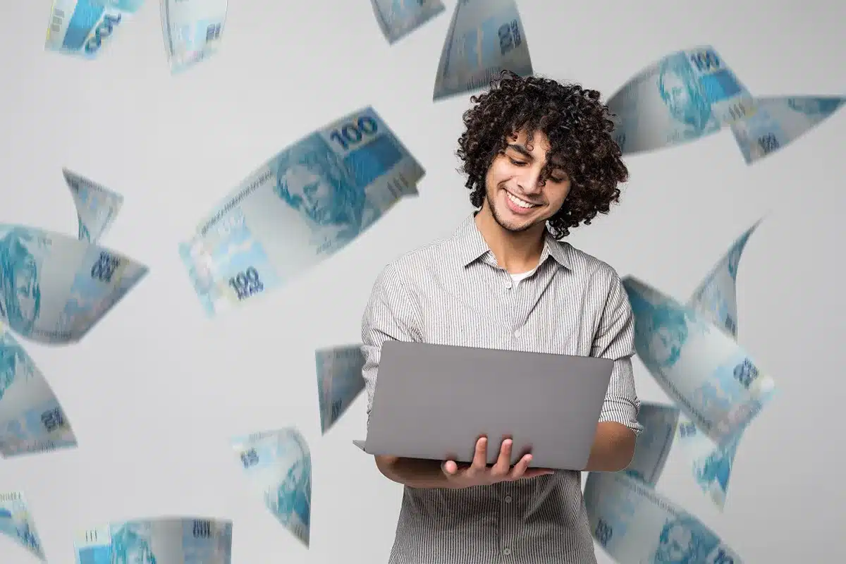 Jovem feliz usando laptop com dinheiro voando ao redor Rapaz sorridente segurando notebook enquanto cédulas de cem reais caem ao redor em fundo cinza