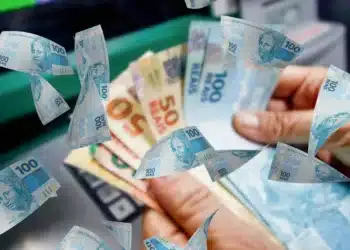 Mãos segurando diversas cédulas de reais com notas de 50 e 100 reais, dinheiro voando ao redor