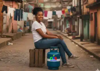 Mulher negra sorridente sentada em caixa de madeira com botijão de gás azul em rua de comunidade com roupas penduradas