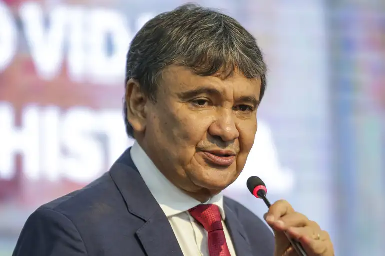 Político discursando em evento formal Homem vestido com terno azul e gravata vermelha falando no microfone em evento ao fundo desfocado