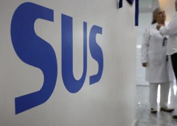 Close na sigla "SUS" em letras azuis sobre uma parede branca de hospital, com profissionais de saúde ao fundo conversando em um corredor iluminado.